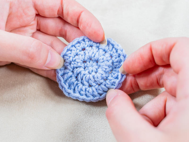 Gros plan de deux mains tenant une petite pièce ronde au crochet bleu clair créée avec un cercle magique au crochet sur un fond de tissu pâle.