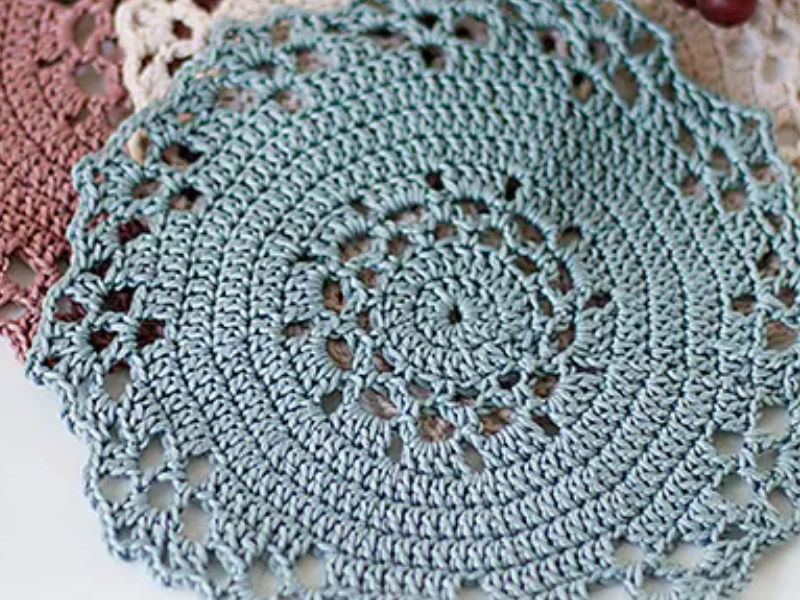 Gros plan d'un napperon rond bleu clair en crochet avec des motifs complexes et des bords festonnés, chevauchant partiellement d'autres napperons dans des tons marron et beige en arrière-plan.