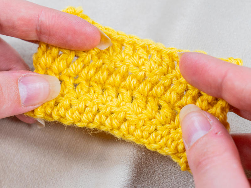 Gros plan de deux mains tenant un petit morceau de tissu crocheté jaune, mettant en valeur la texture, les détails de la mariée au crochet et les points clairement sur un fond beige clair.