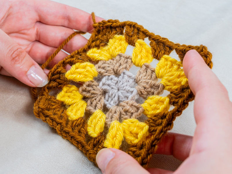 Gros plan sur des mains tenant un carré en crochet coloré avec des fils jaunes, bruns et beiges, sur un fond de tissu clair.