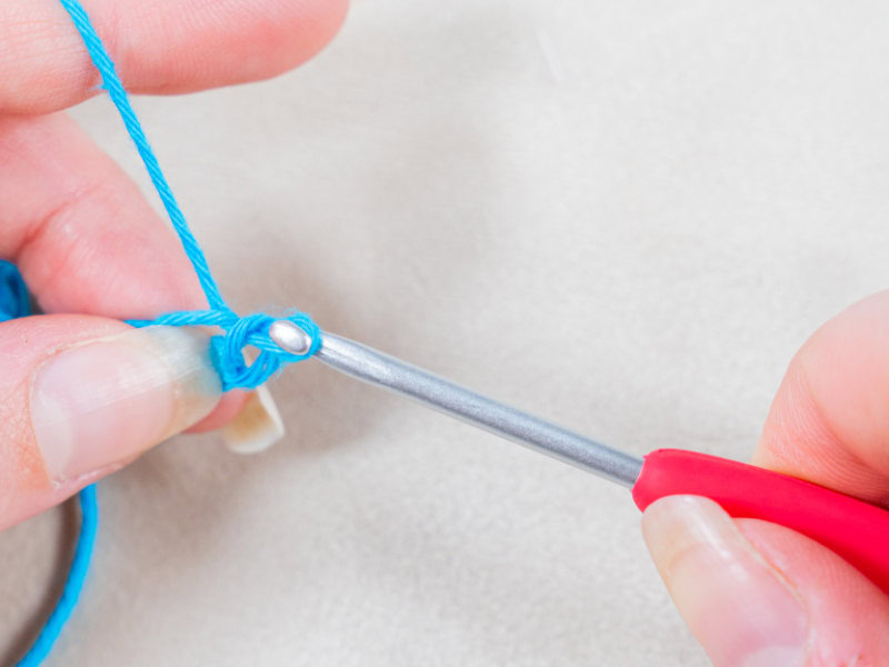 Gros plan sur des mains tenant un crochet à manche rouge et un fil bleu, formant une maille sur un fond clair - parfait pour illustrer le crochet en anglais.