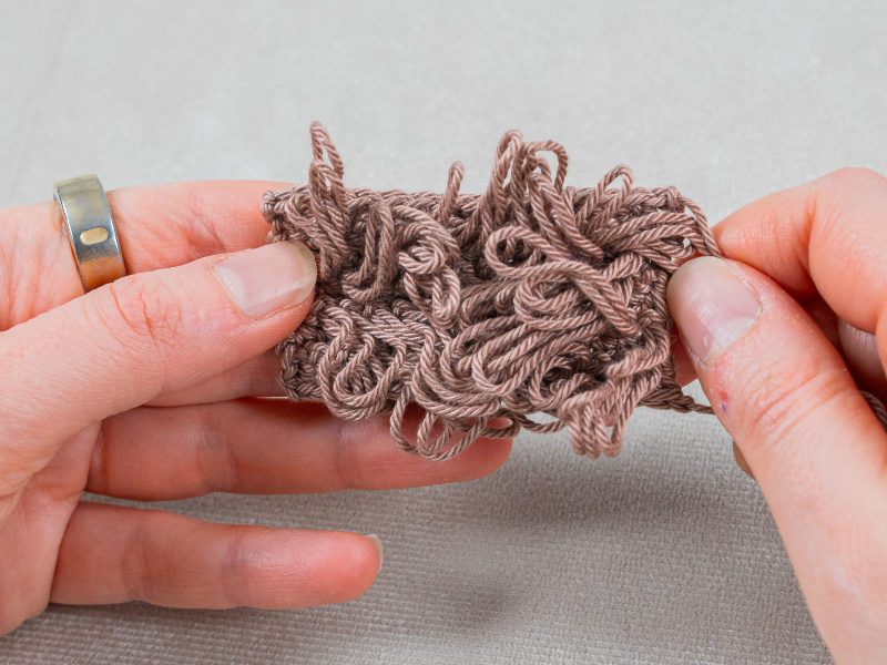 Deux mains tiennent un morceau de fil brun et frisé travaillé au point de crochet, mettant en valeur son aspect texturé et enchevêtré sur un fond neutre.