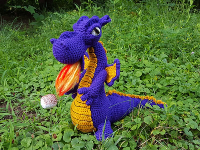 Un dragon au crochet en violet et jaune, avec une bouche ouverte et des flammes, est assis sur de l'herbe verte et des plantes feuillues à l'extérieur.