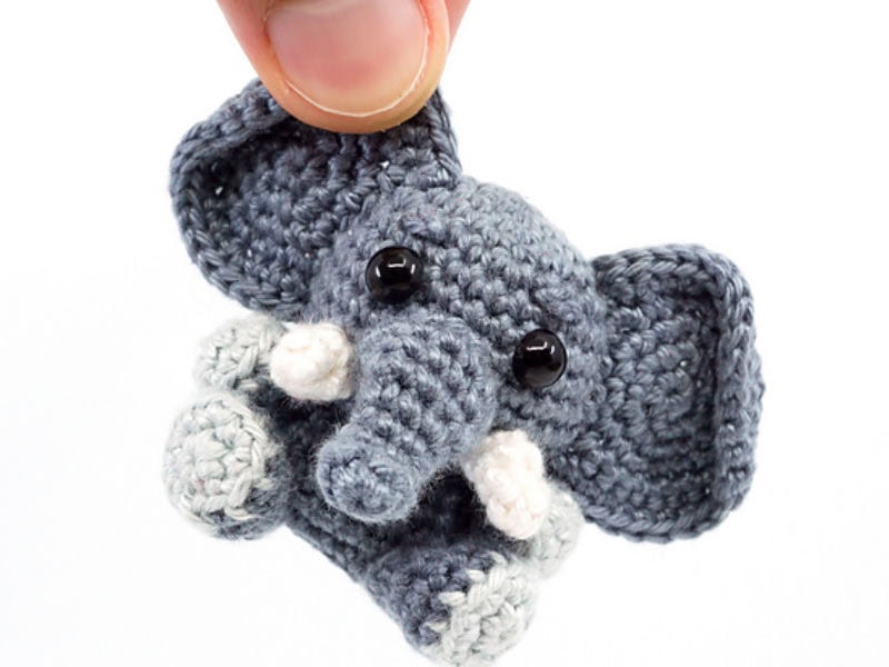 Une main brandit un petit éléphant au crochet avec de grandes oreilles, une longue trompe, des yeux noirs en forme de boutons et des défenses blanches.
