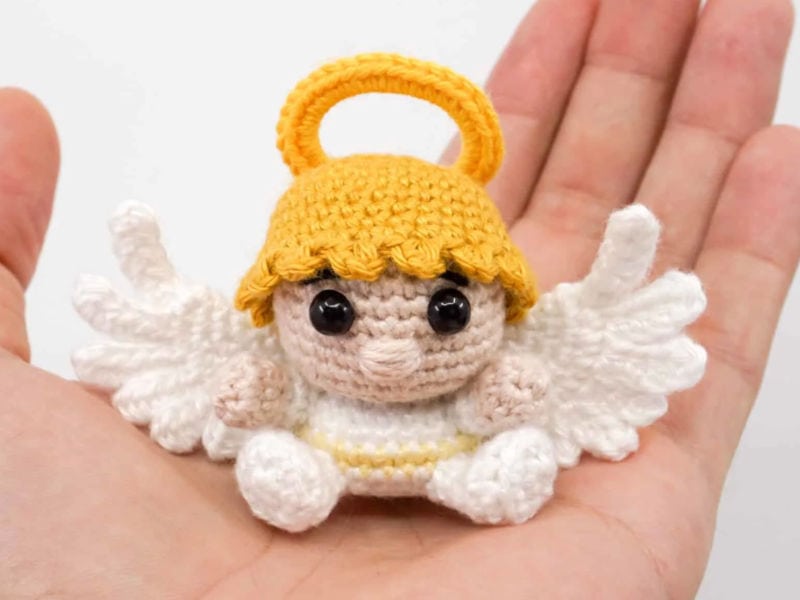 Un petit ange au crochet, aux ailes blanches délicates et à l'auréole jaune, est posé dans la paume d'une main, sur un fond uni.