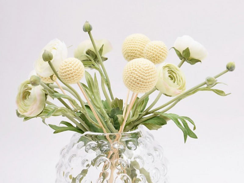 Un vase en verre transparent, dont la surface est texturée et présente un aspect alvéolé, contient un bouquet de fleurs au crochet dans des tons jaune pâle et blanc, dont certaines ont la forme de boules crochetées, agrémenté de feuilles et de bourgeons verts.
