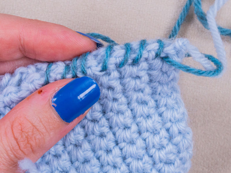 Une main aux ongles vernis de bleu tient un tissu au crochet bleu clair, tandis qu'un fil bleu-vert est cousu sur le bord supérieur, mettant en valeur un travail de haute couture au crochet d'une grande finesse.