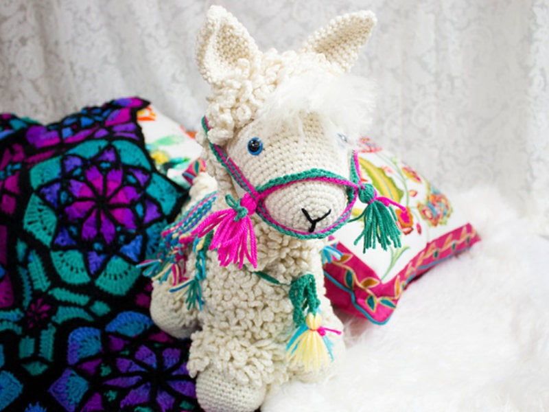 Un lama au crochet, orné de pompons colorés sur son harnais, est posé sur une surface blanche, à côté de coussins aux motifs éclatants et d'une couverture au crochet.