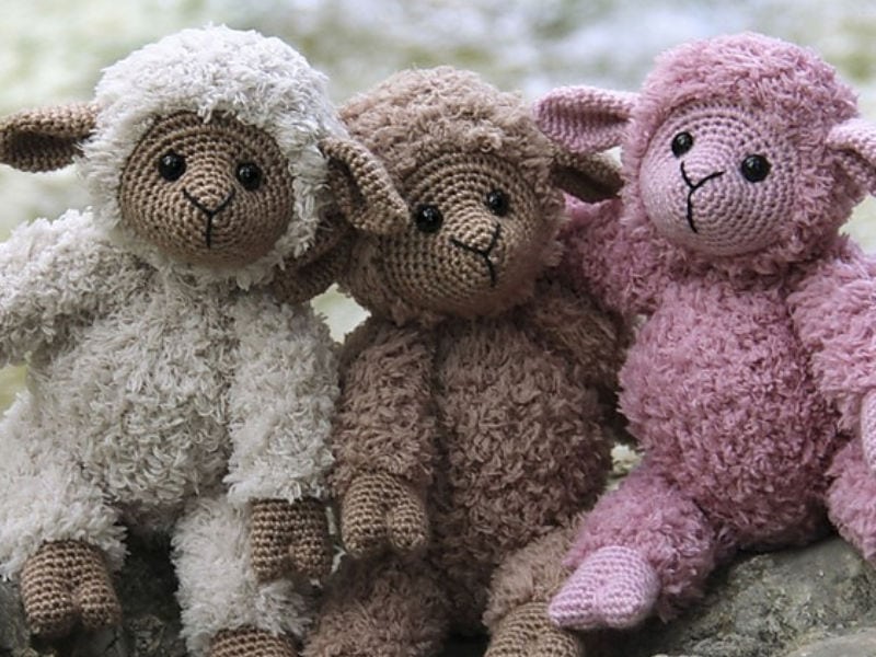 Trois peluches au crochet représentant des moutons sont assises côte à côte. Ces charmants « moutons au crochet » sont blancs, marron et roses ; chacun a des yeux en boutons et une fourrure douce et texturée, et ils sont représentés assis près les uns des autres, l’air doux. L’arrière-plan est flou et naturel.