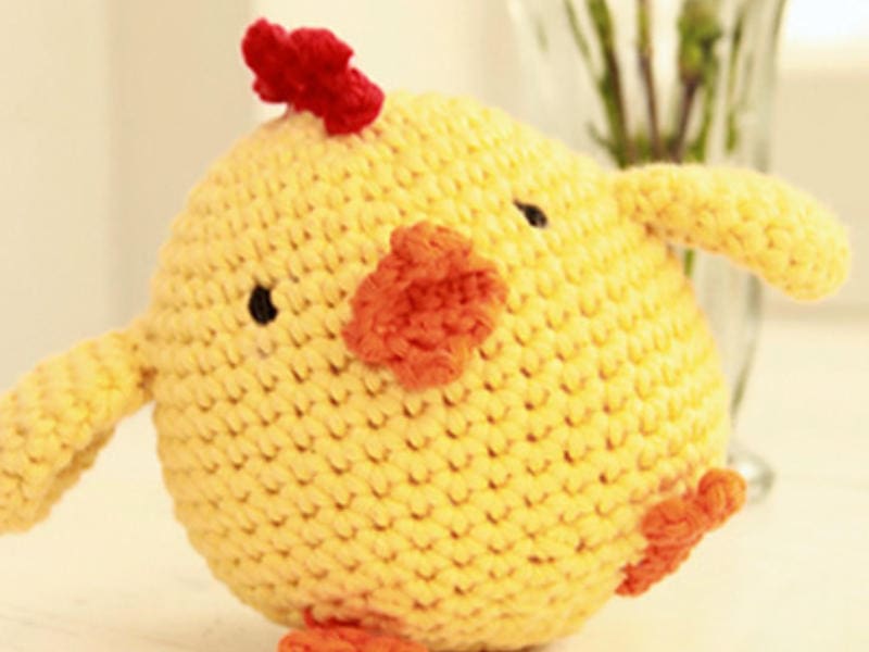 Un poulet rond et jaune, réalisé au crochet — idéal pour toute collection de « poules au crochet » —, doté d'un bec et de pattes orange, d'yeux noirs brodés et d'une crête rouge, est posé sur une surface blanche, avec en arrière-plan un vase en verre aux contours flous.