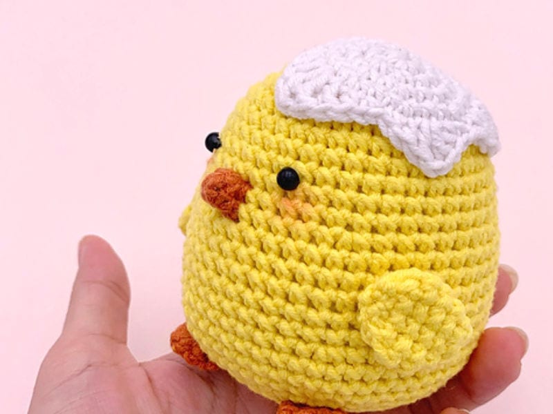 Une main tient un adorable poussin au crochet — un poussin jaune au petit bec orange, aux ailes minuscules, aux yeux noirs et coiffé d'une coquille d'œuf blanche au crochet — sur fond rose pâle.